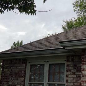 A view of a brick house corner with a brown shingled roof, rain gutter, and windows against a cloudy sky.
