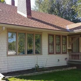 The exterior side of a house with light-colored siding, a brown shingled roof, a chimney, and several large windows.