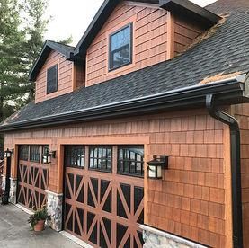 A two-car garage with cedar shingle siding, carriage-style doors with glass windows, and black gutters.