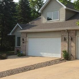 Tan brick and beige siding house with a white two-car garage door and a gravel strip along a paved driveway.
