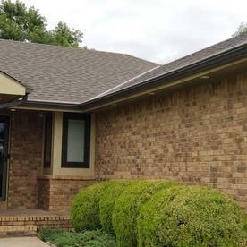 A tan brick house exterior with a grey shingled roof, dark window frames, and round green shrubs in the front yard.