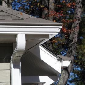 A close-up of white aluminum rain gutters and a downspout attached to the corner of a house with beige siding.