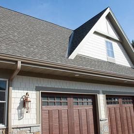 A tan house with a gray shingled roof, a front-facing gable, and two wooden garage doors with windows.