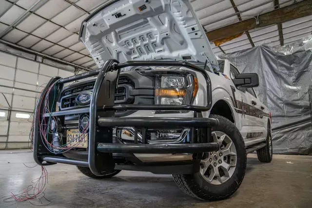 White Ford pickup truck with hood open, in a garage. Black grill guard and license plate visible.