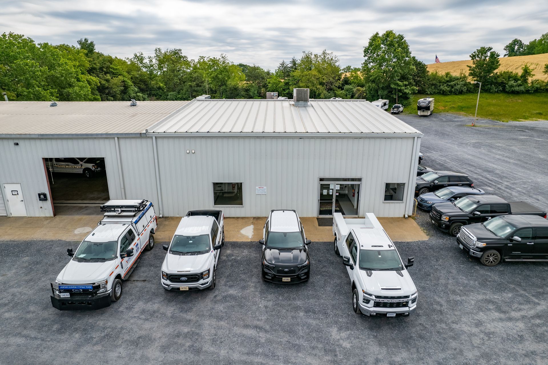 Vehicles parked in front of a white building with trees in the background. Gravel lot. Cloudy sky.