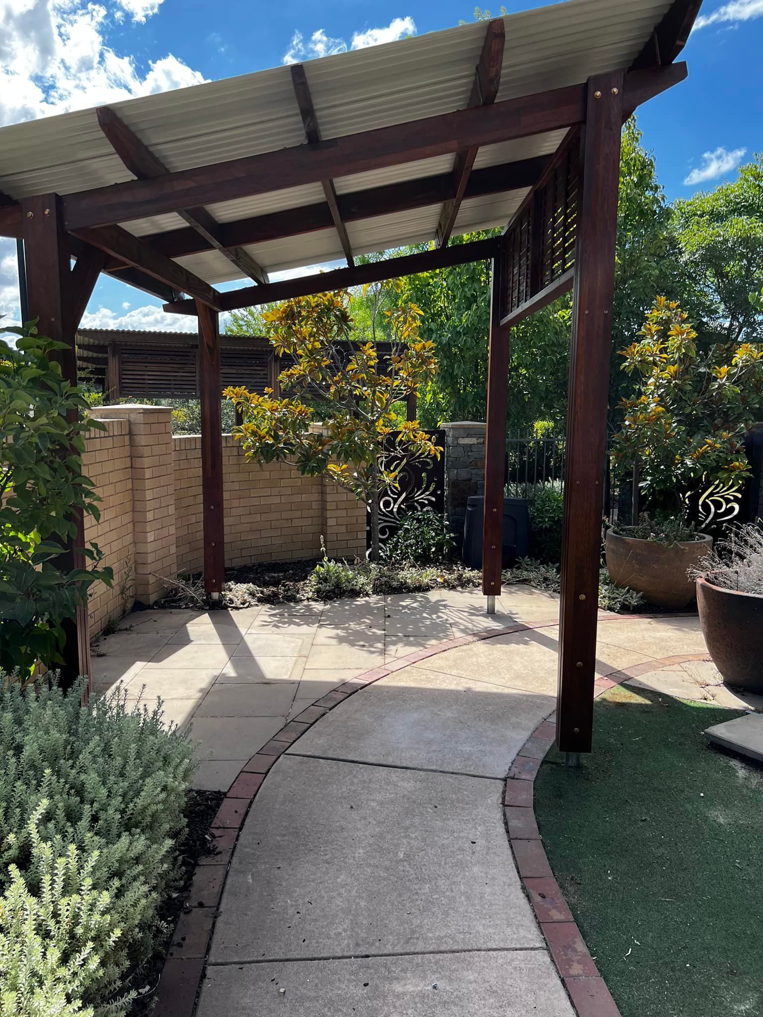 A Wooden Pergola with A Metal Roof Is in The Middle of A Garden — P & M Maintenance Building Services in Yass River, NSW