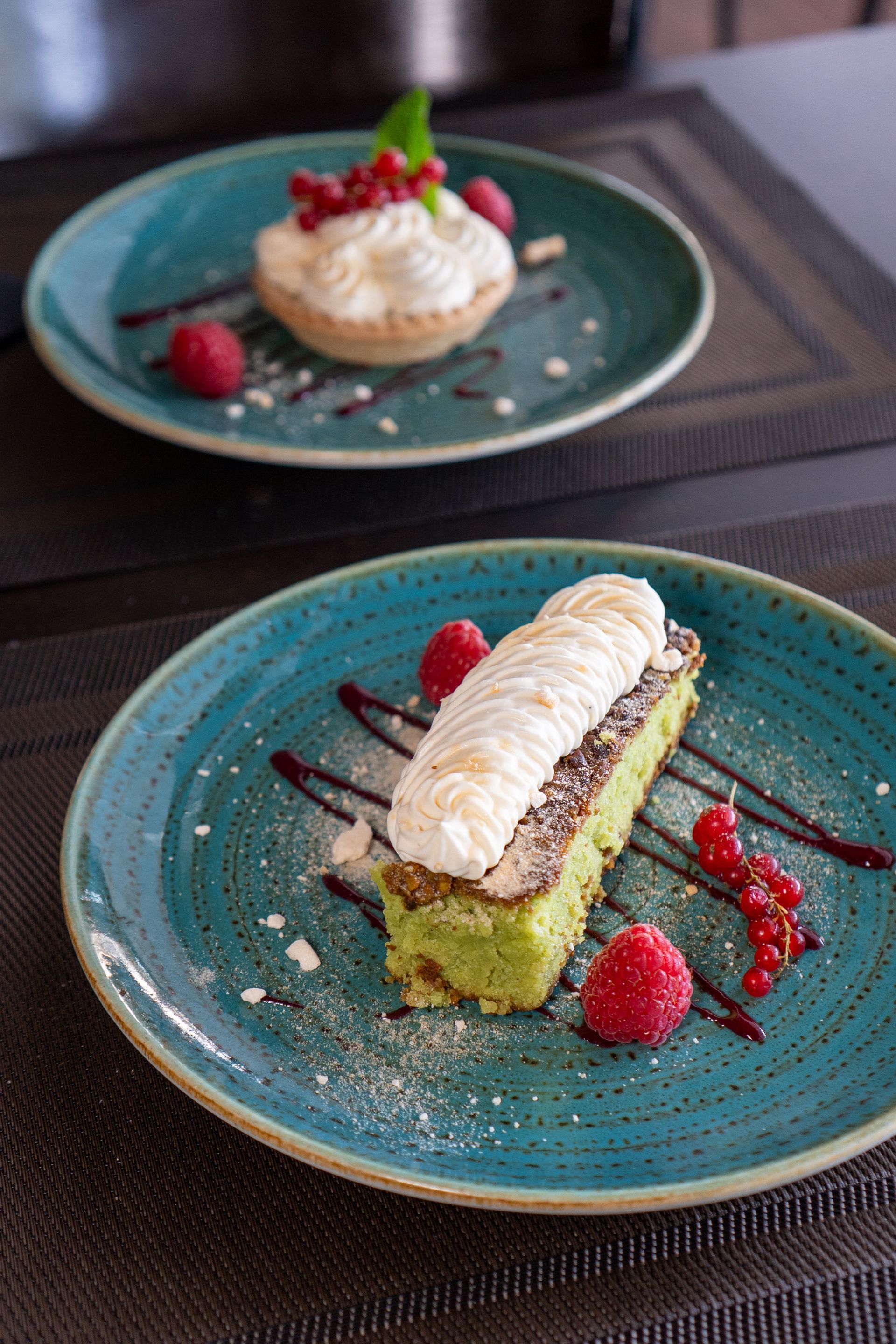 Deux desserts sur des assiettes bleu sarcelle, garnis de crème, de baies et de sauce, sur une table.