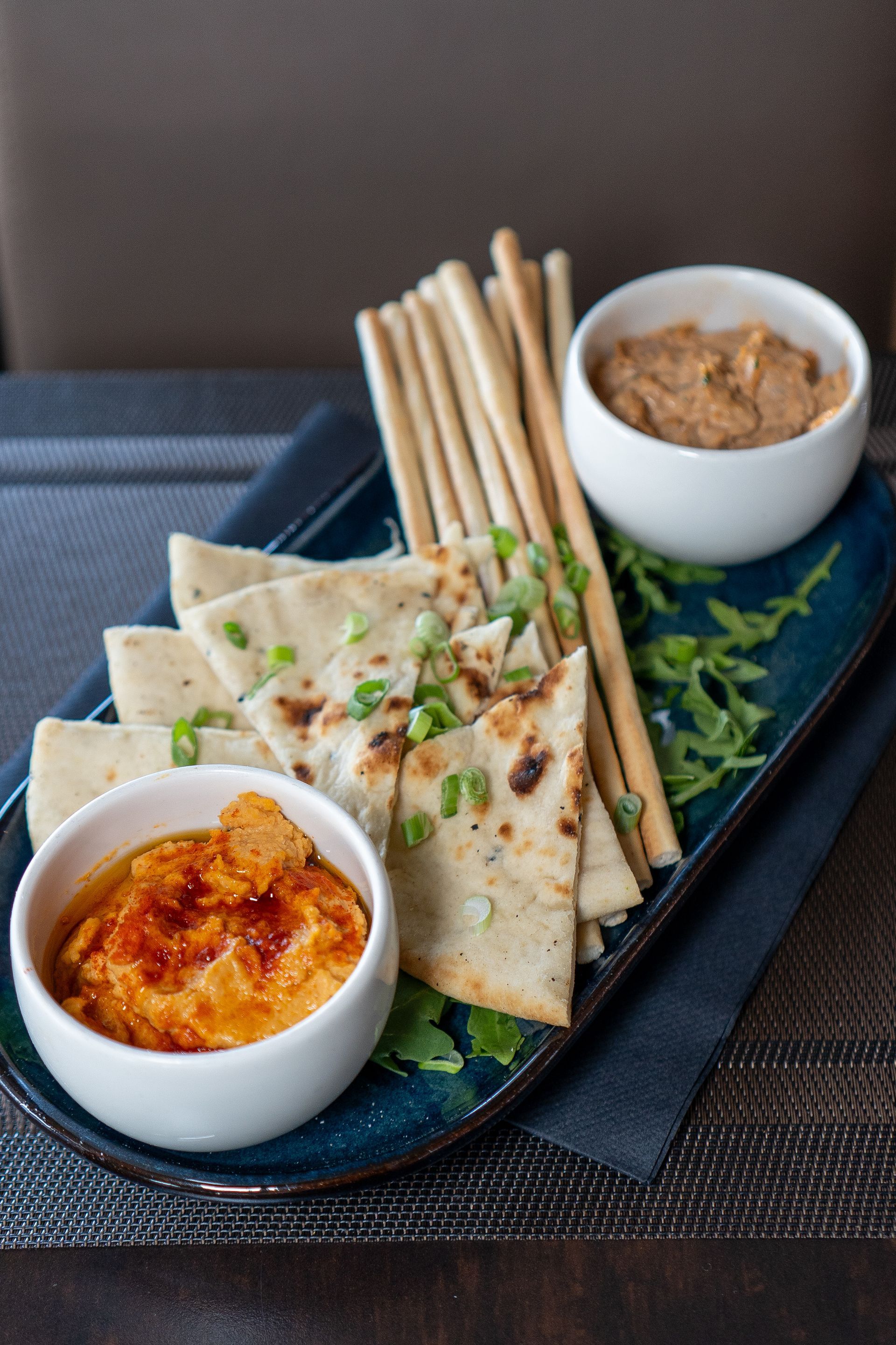 Apéritif au houmous : pain pita, gressins, deux bols blancs de houmous et garniture verte sur un plateau bleu.