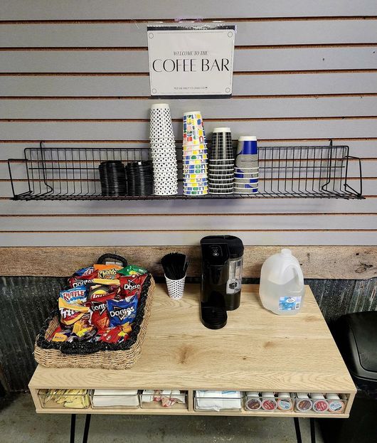 Coffee station with cups, mugs, snacks, and a water jug on a wooden counter against a slat wall. | Dillon's Automotive
