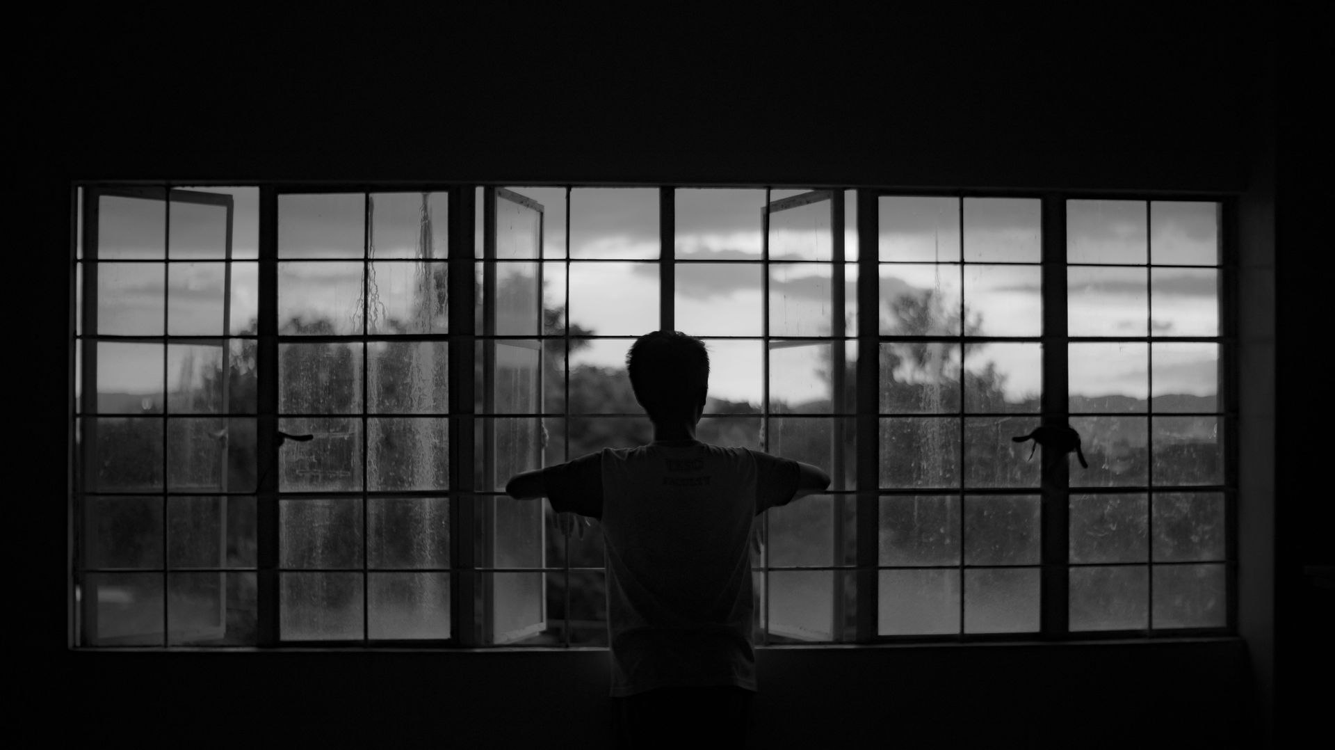 A photograph of someone sitting alone by a window at dusk, back partly turned to camera.