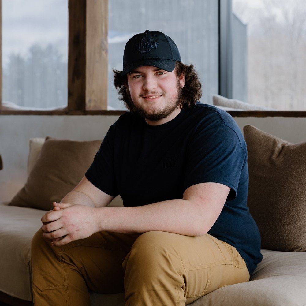 Man in a black cap and shirt sitting on a couch, smiling. He is in a room with natural light.