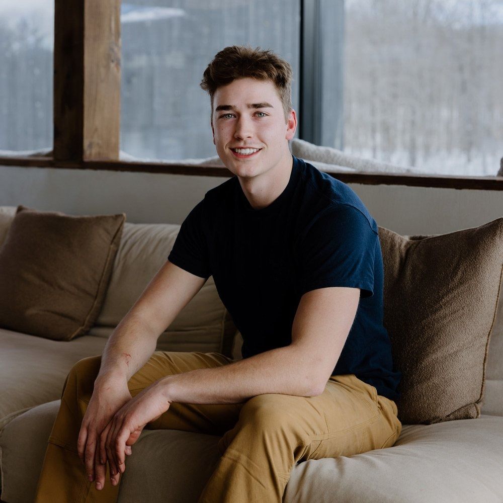 Young man smiles, sitting on a couch indoors. He wears a navy shirt and tan pants.