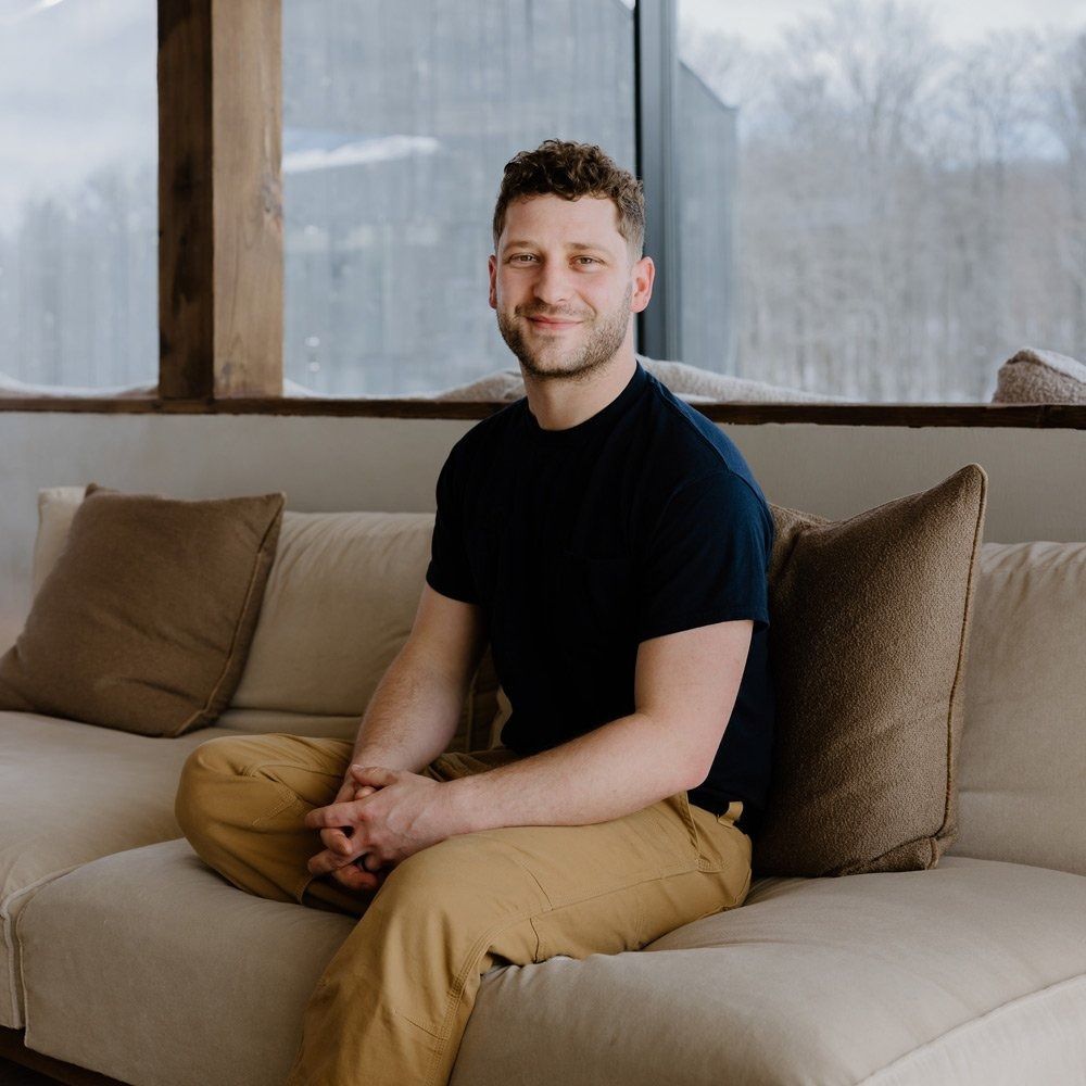Man sitting on a light sofa, wearing a navy shirt and tan pants, smiling. Indoors, natural light.