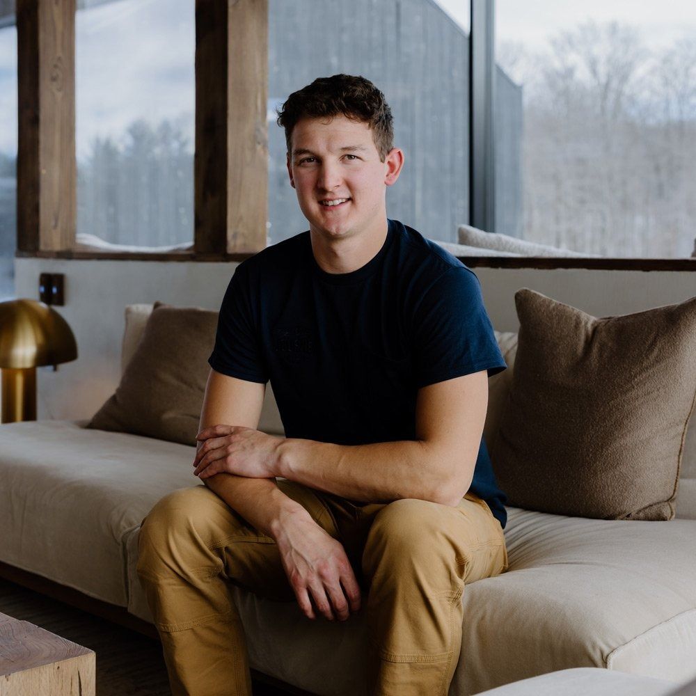 Man sitting on a couch, smiling; wearing a navy shirt and khaki pants. Modern interior with a window background.