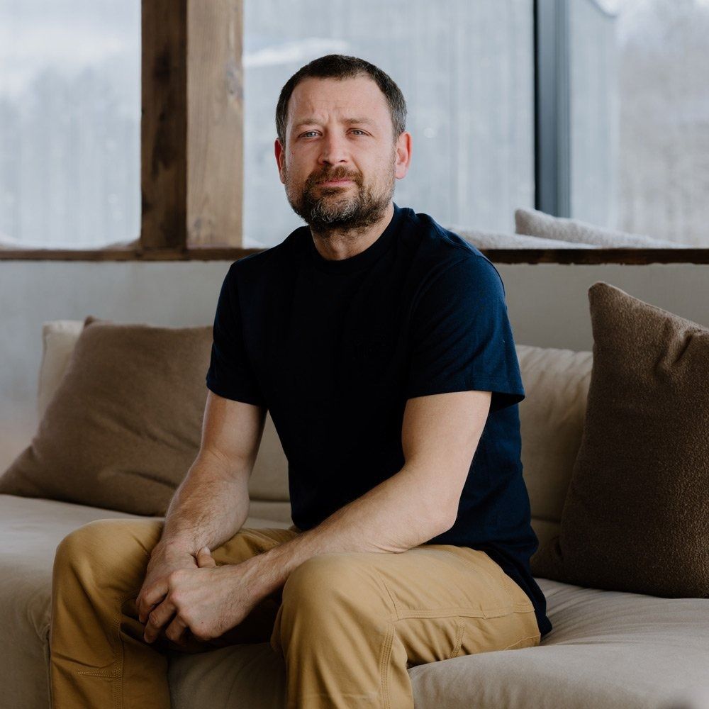 Man with beard sitting on a cream couch, wearing a navy t-shirt and khaki pants.