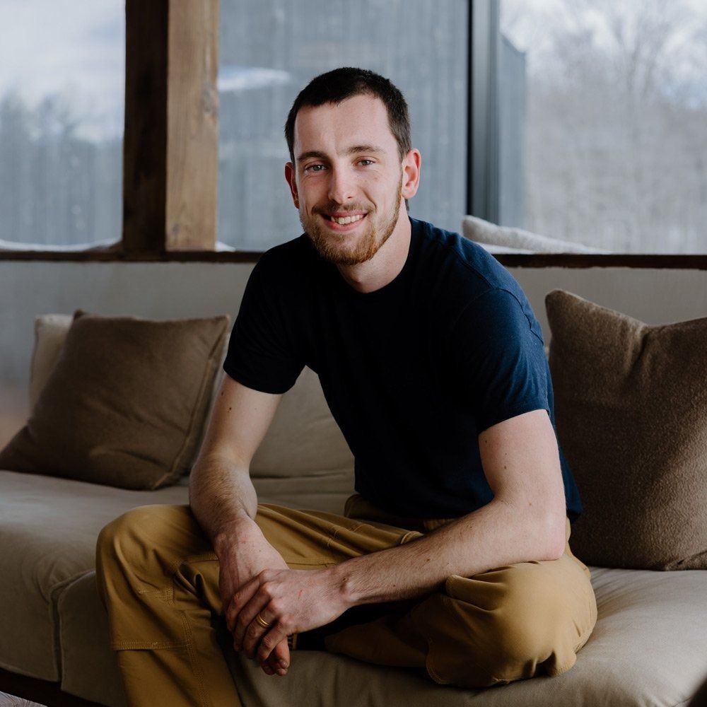 Man with a beard smiles, sitting on a couch. He wears a navy shirt and khaki pants indoors, with large windows behind him.