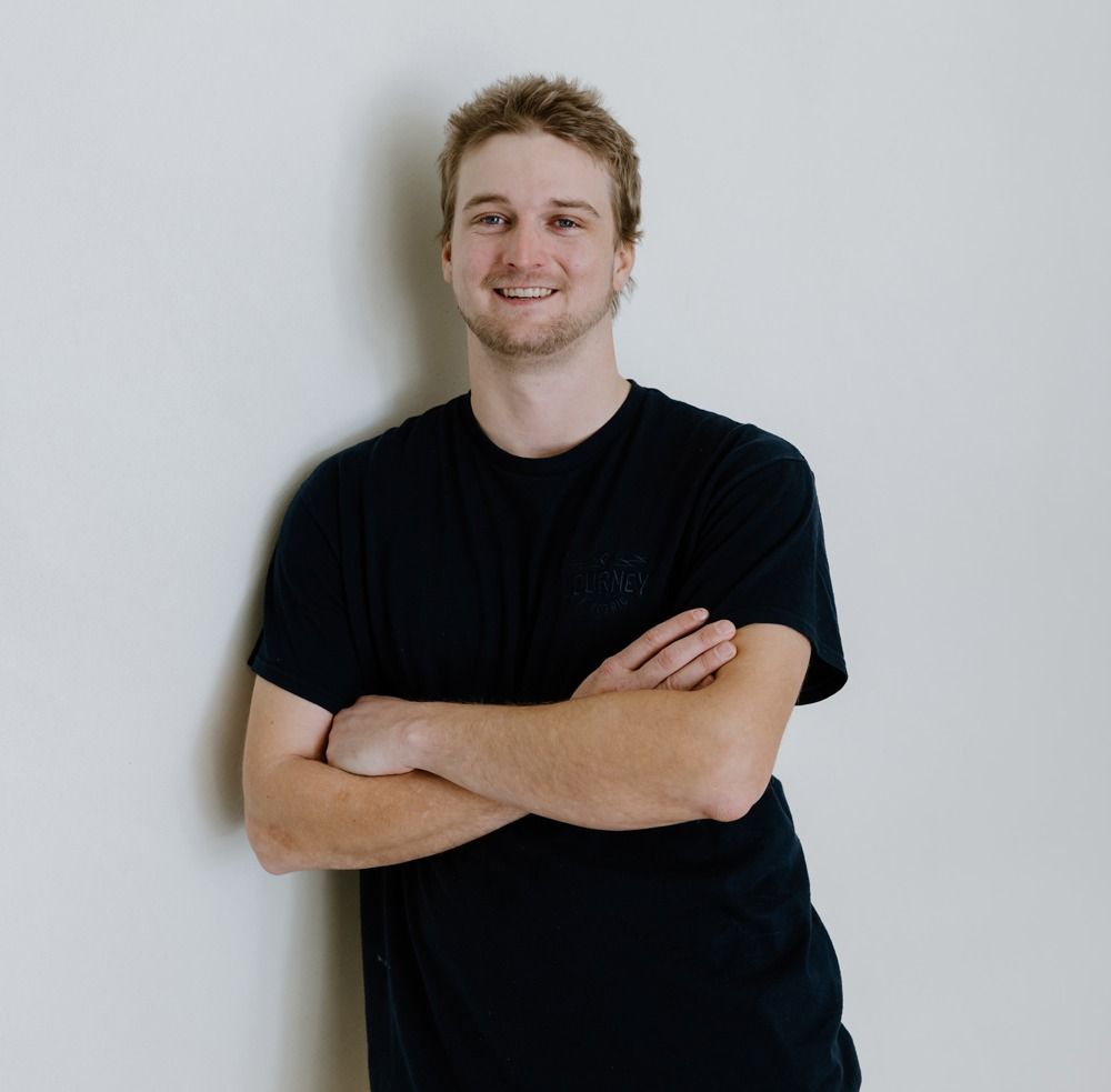 A young man with blonde hair smiles, arms crossed, leaning against a white wall. He wears a black t-shirt.