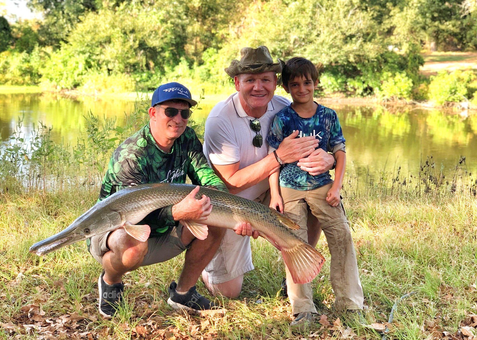 Three people with fish by water. Men and a boy smiling, holding large fish on grass.