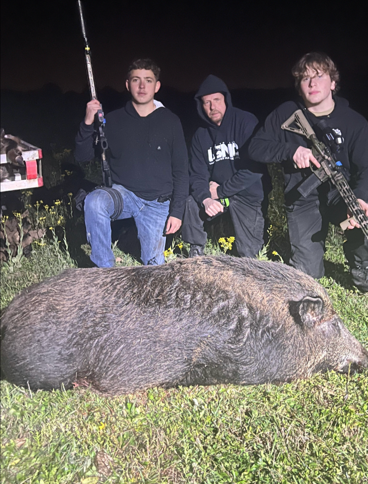 Three men pose with a large dead wild pig at night; one kneels holding a rifle, others squat.