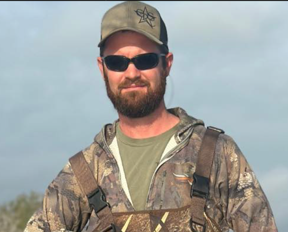 A bearded person wearing dark sunglasses, a camouflage cap, and a camouflage jacket outdoors under a cloudy sky.