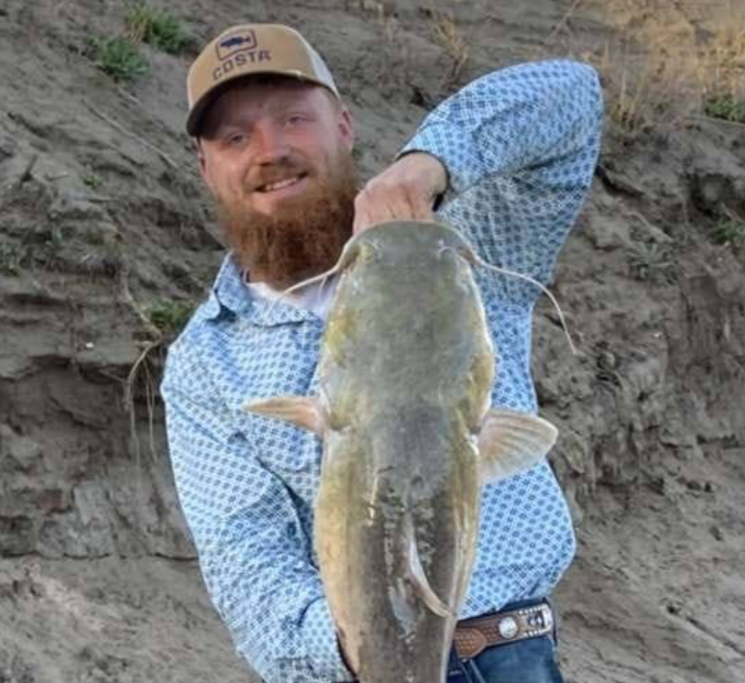 A bearded person in a blue patterned shirt and baseball cap smiling while holding a large catfish against a dirt bank.