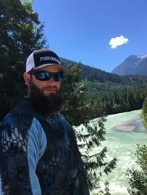 A bearded person wearing a fishing cap and sunglasses, standing in front of a turquoise river and a forested mountain.