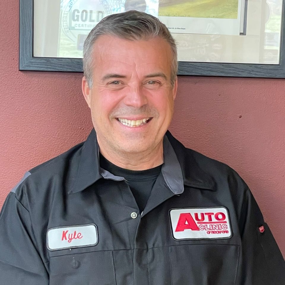 Smiling auto service employee in black shirt with name tag, standing in front of a wall.