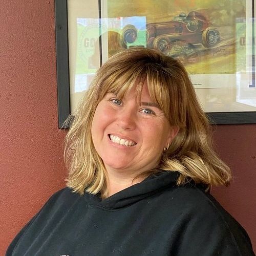 Smiling person in a black hoodie standing indoors against a red wall with framed artwork.