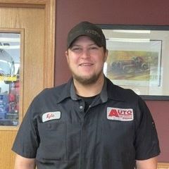 Smiling auto shop employee in a black uniform and cap standing indoors near framed wall art.