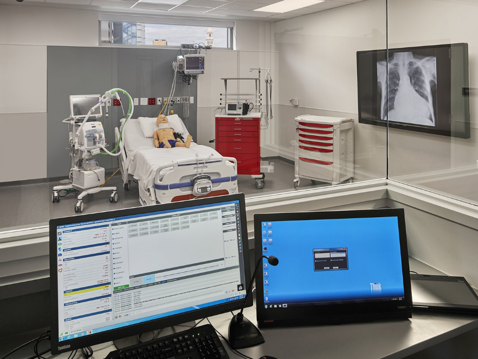 A computer monitor is sitting on a desk in front of a hospital bed.
