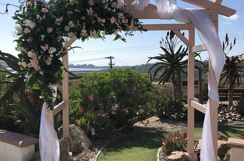 Floral archway for an outdoor event with white fabric draped, overlooking greenery and a body of water.