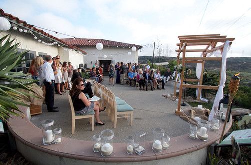 Wedding ceremony outside; guests gathered; wooden arch with fabric; candles on a curved wall; white building.