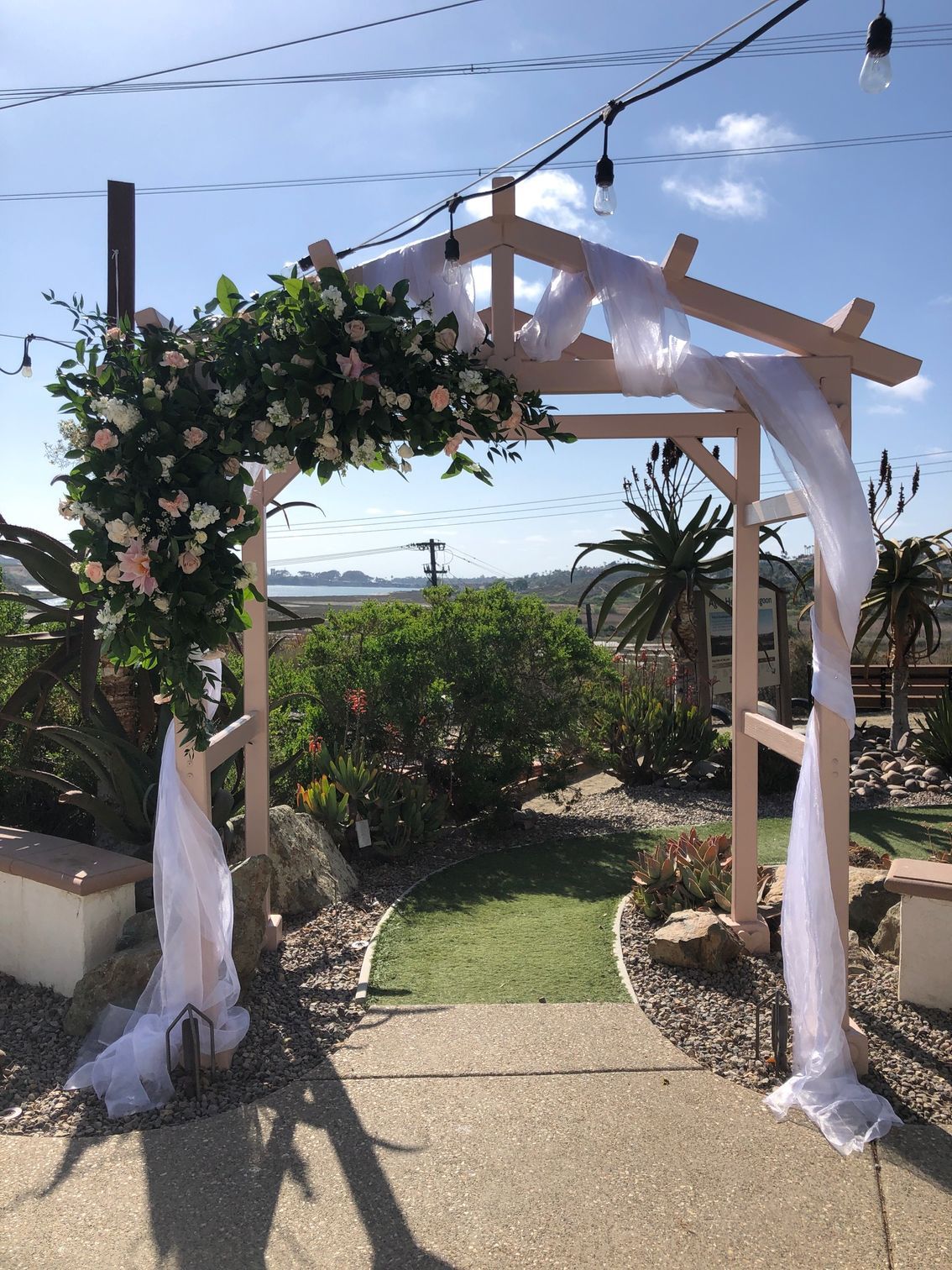 Wooden wedding archway draped with white fabric and flowers, overlooking a green yard and water view.