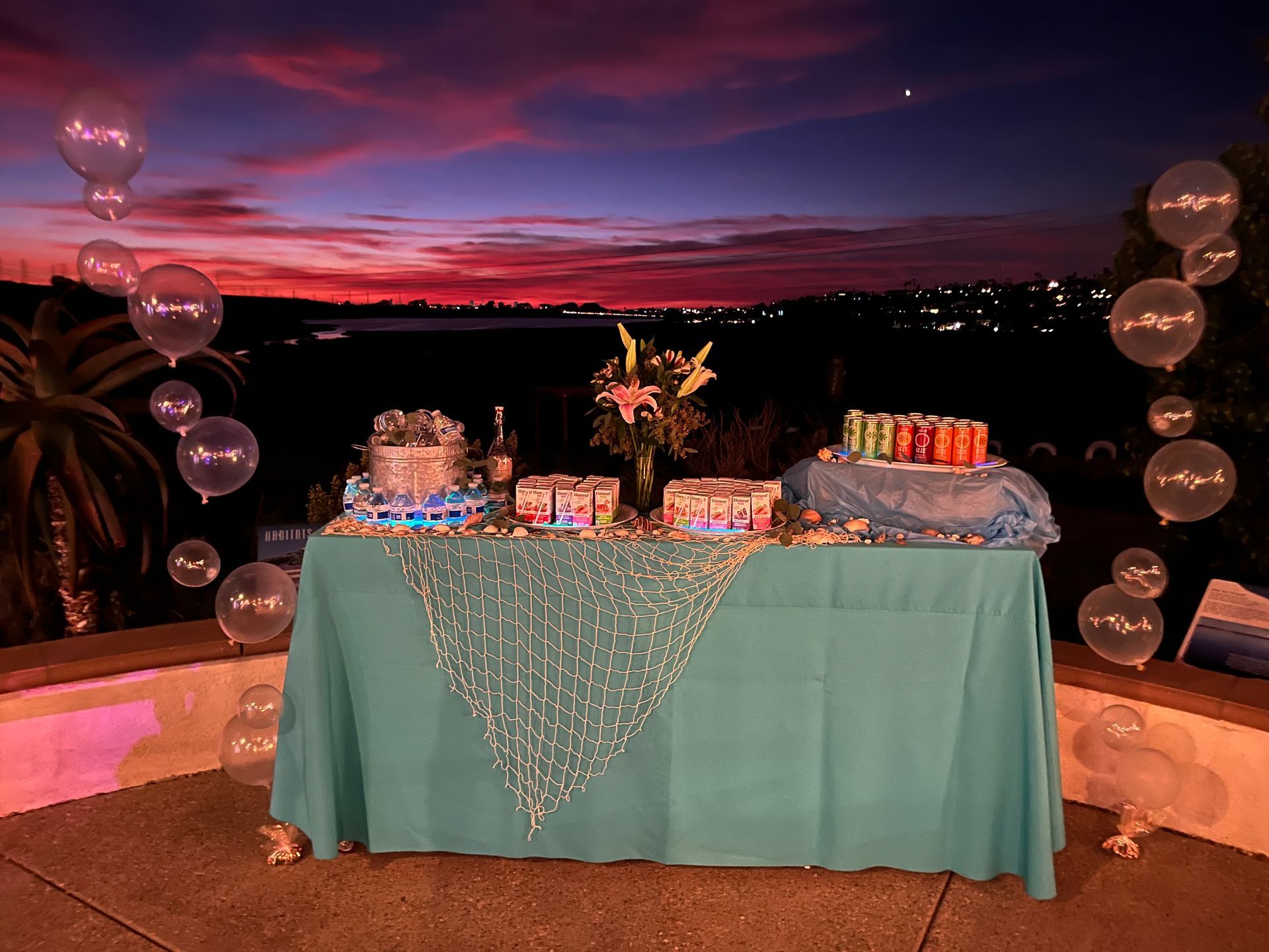 A teal party table with snacks and drinks, decorated with balloons against a sunset backdrop.