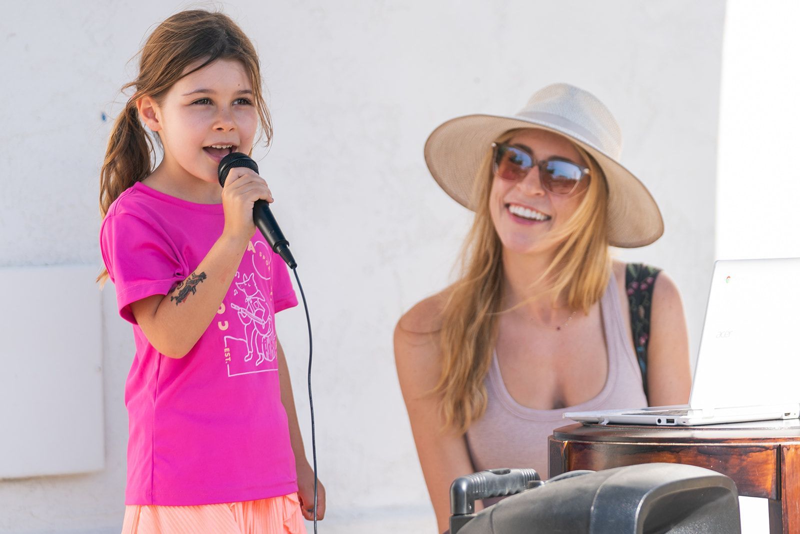 Young girl singing into a microphone beside a smiling woman in a sun hat outdoors