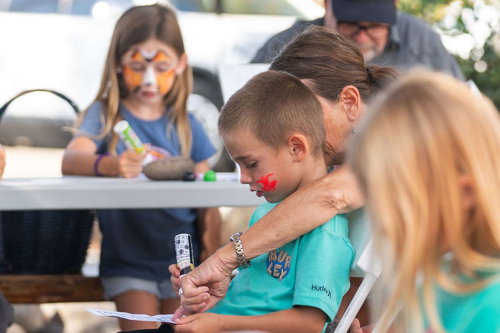 Man helping a child paint at a table while other children work in the background