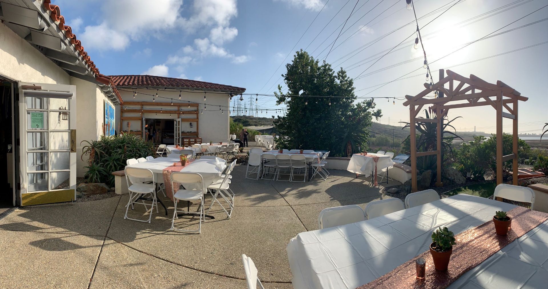 Outdoor patio with tables set for a gathering, strings of lights, a wooden arch, and a building under a cloudy sky.