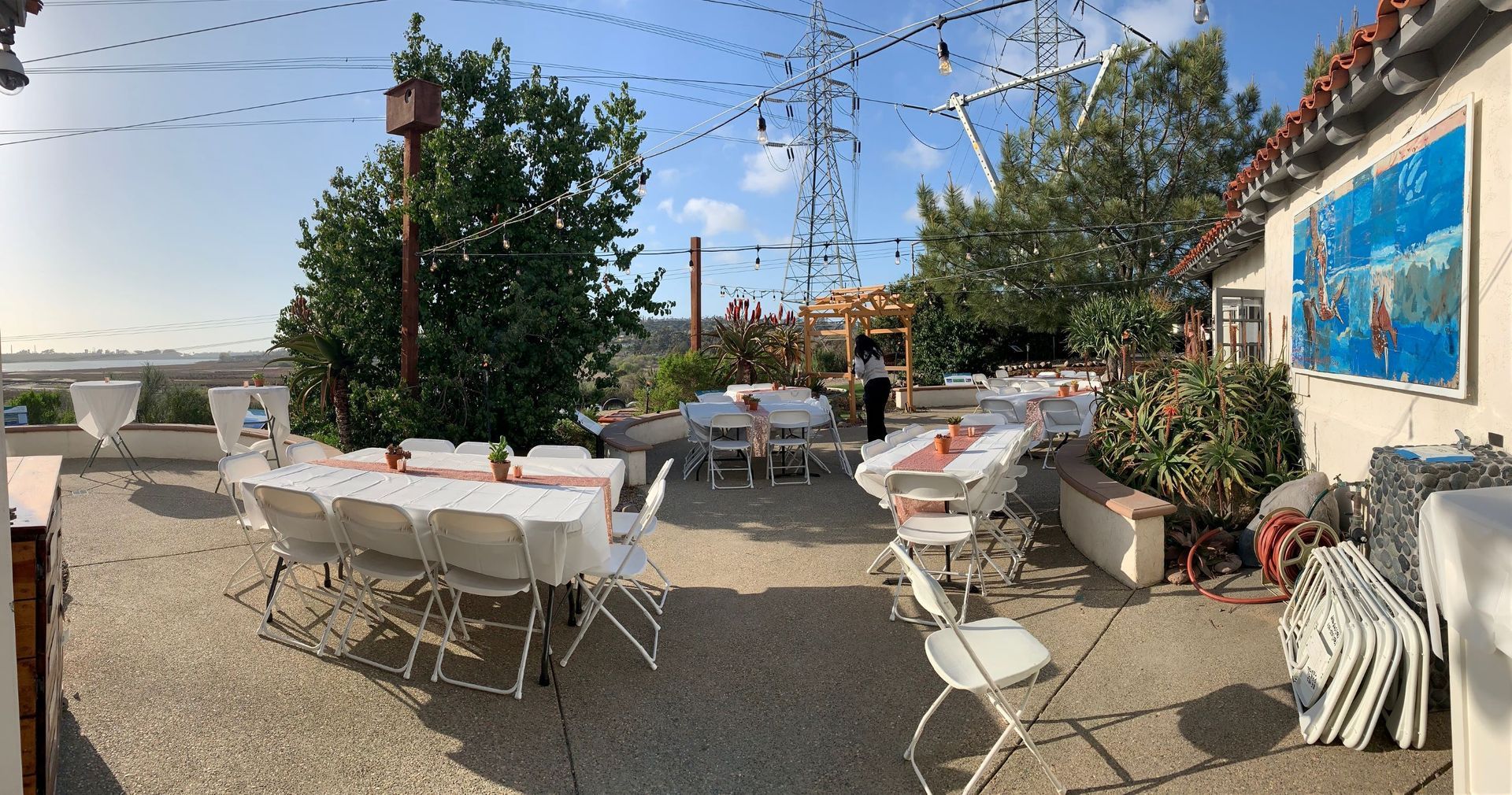 Outdoor event space with white tables set for a party, strung lights, and a mural on the building.