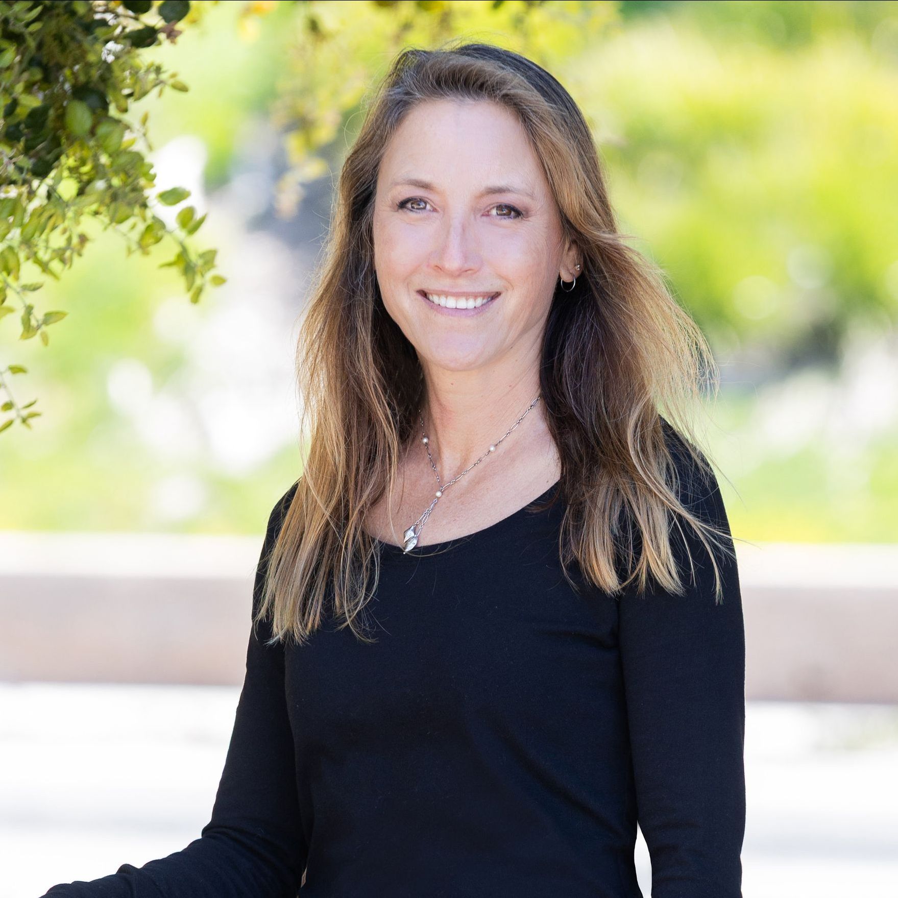 Woman smiling outdoors, wearing a black long-sleeve shirt and silver necklace.