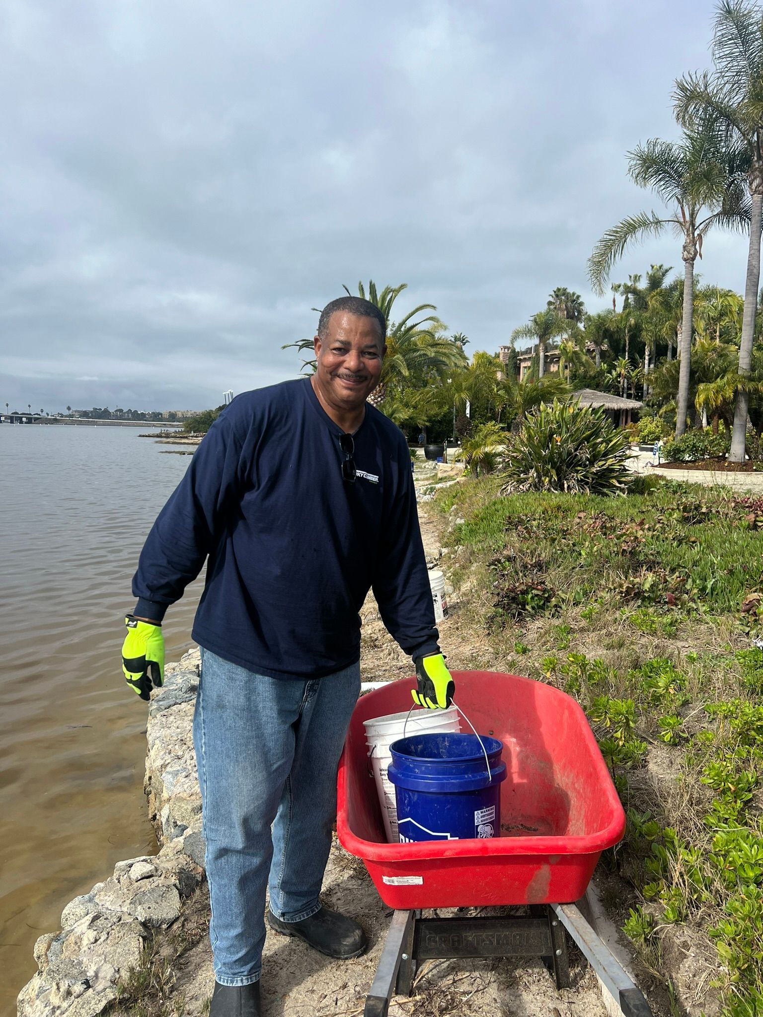 Man smiling, with wheelbarrow containing bucket, near water's edge, wearing gloves, holding wheelbarrow.