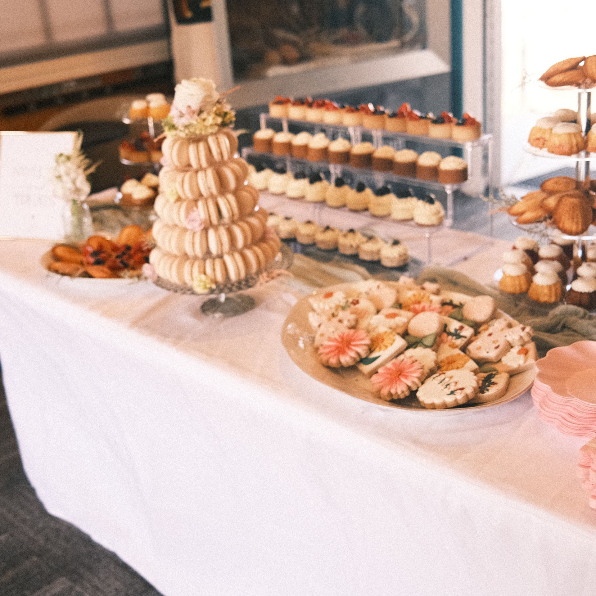 Dessert table with tiered macarons, cupcakes, cookies, and pastries on white tablecloth.