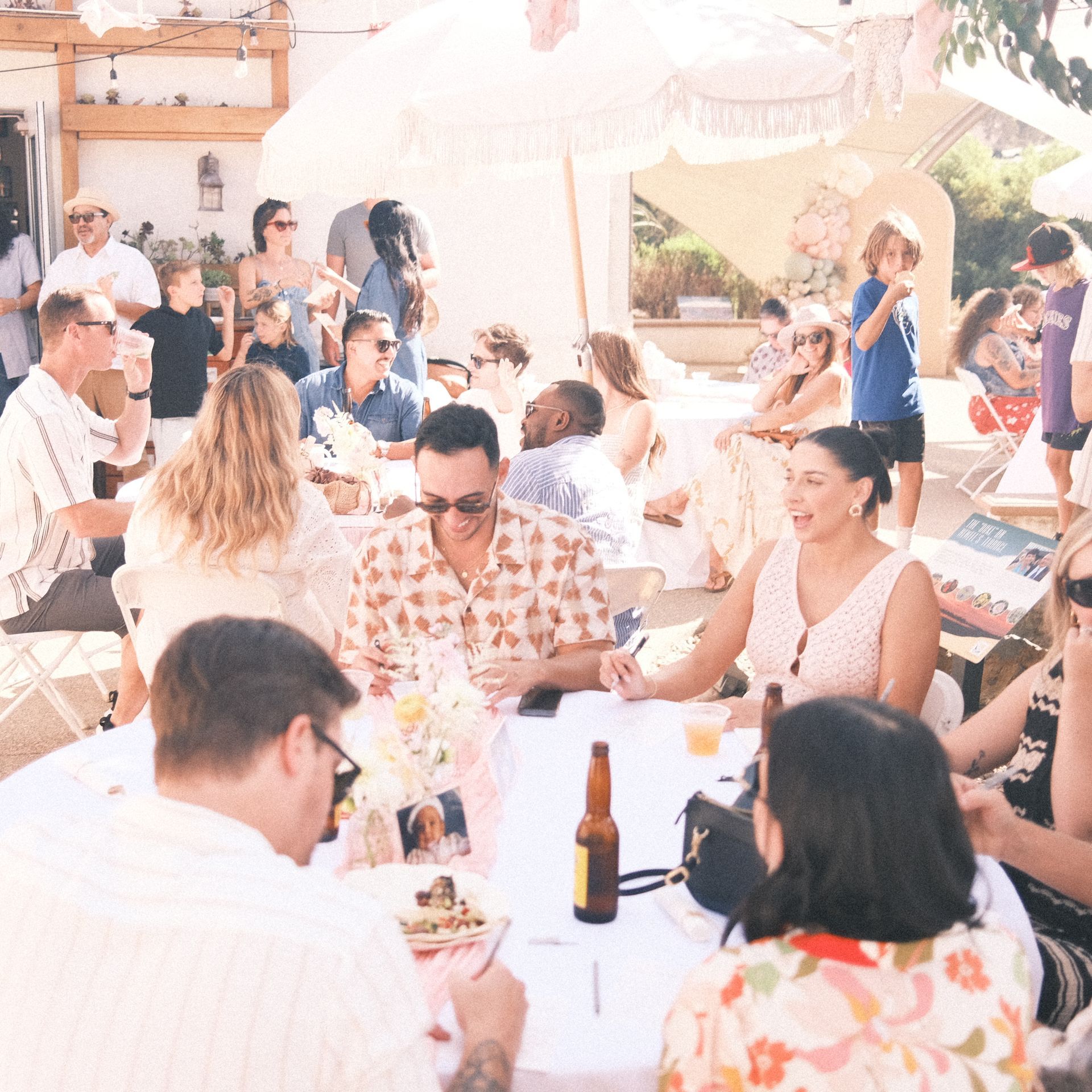People gathered outdoors, sitting at tables, likely celebrating an event. Sunny, relaxed atmosphere.