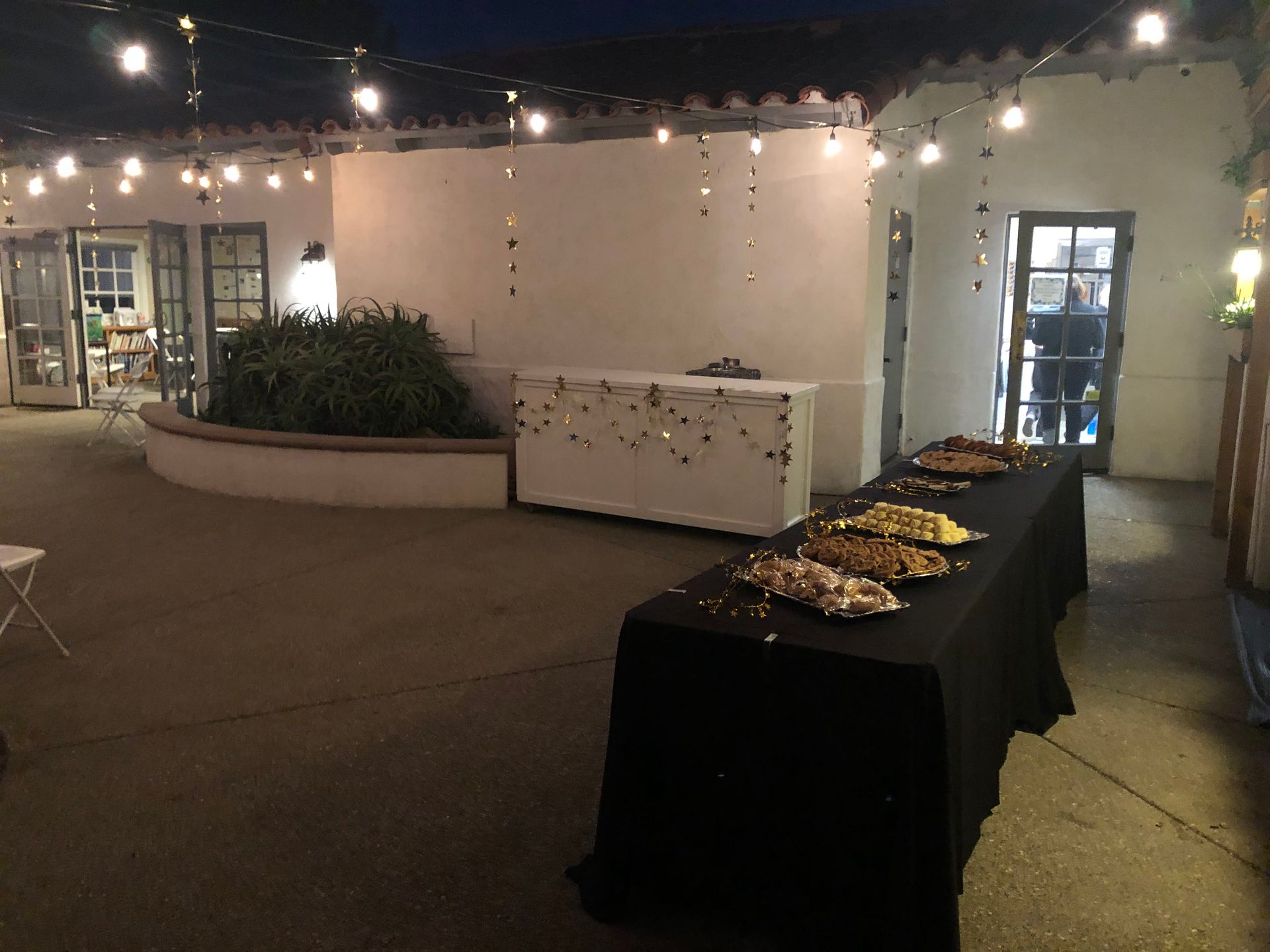 Outdoor event with food on a table, string lights, and white buildings.