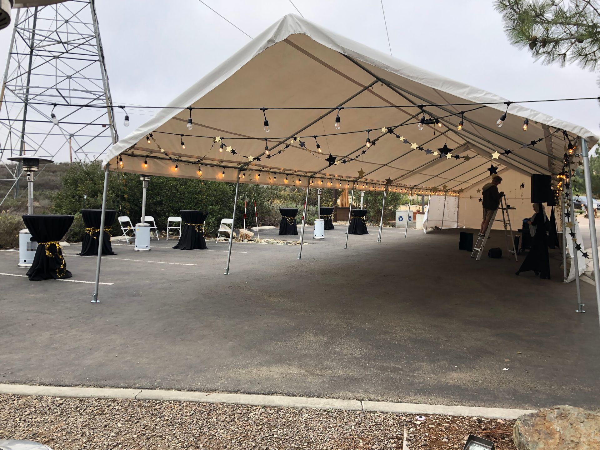 A tent set up for an event, with string lights, tables, and a person on a ladder.