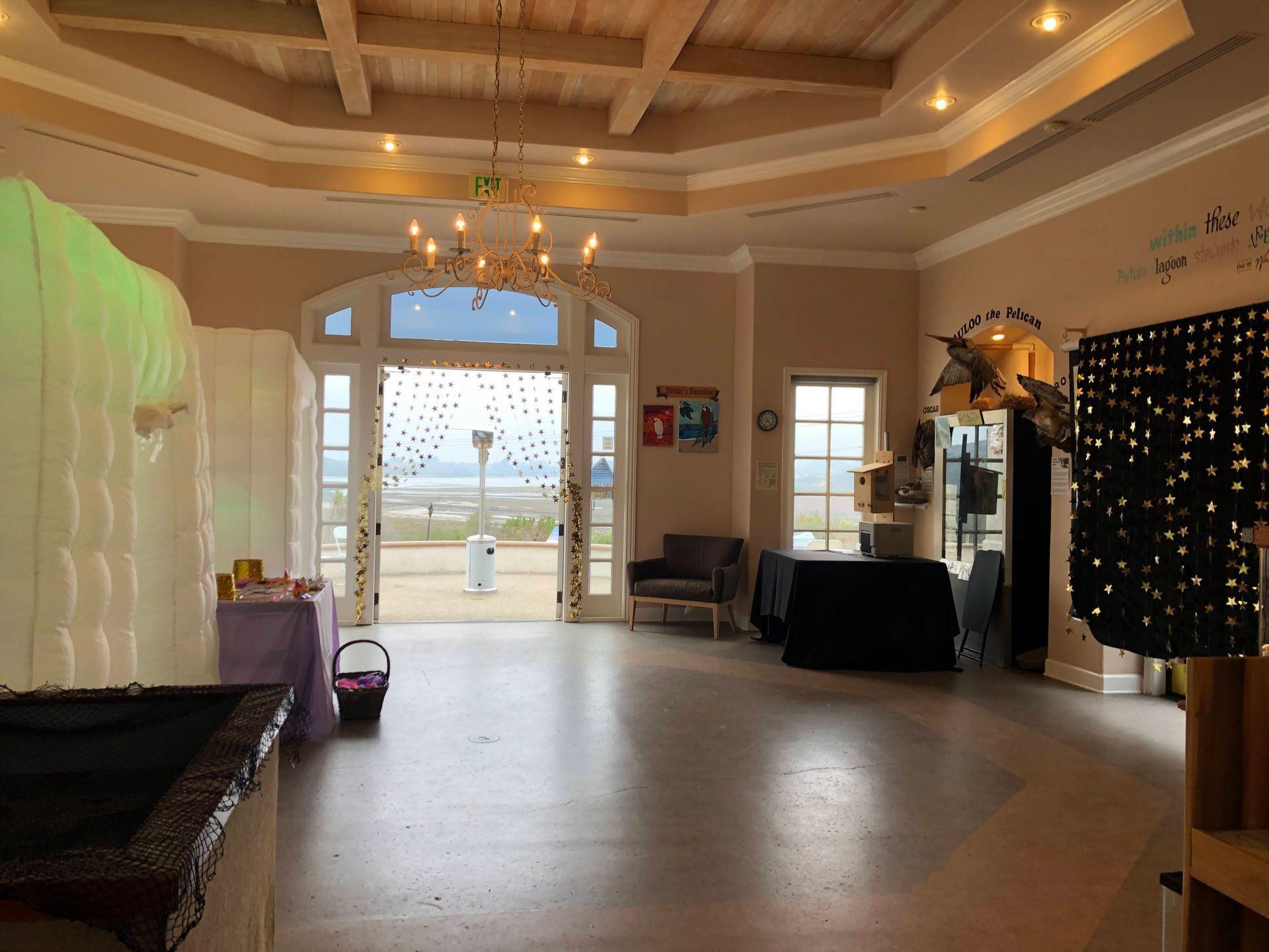 Spacious event hall with open doorway to ocean view. Beige walls, wood ceiling, chandelier, tables, and decorations.