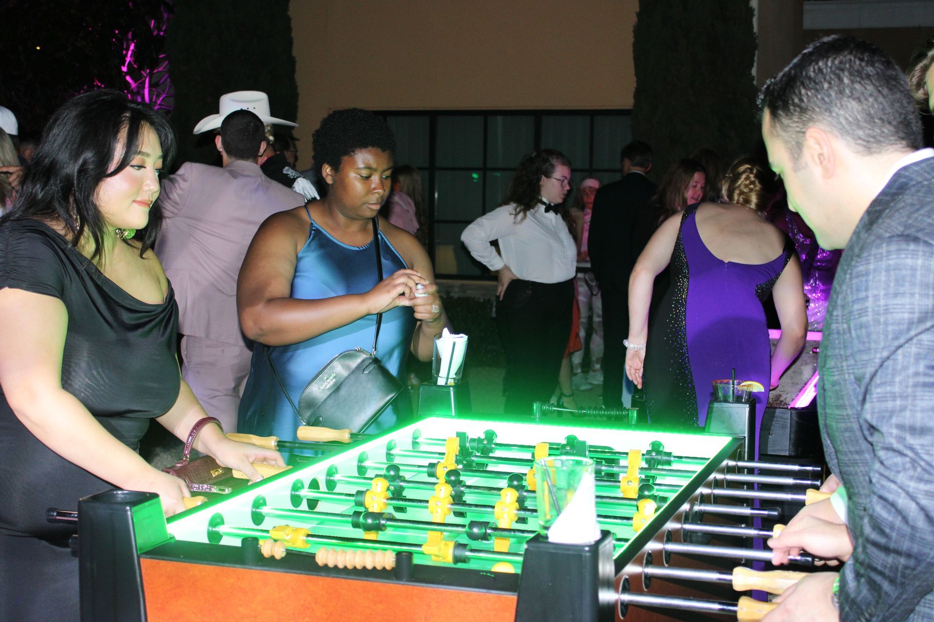 People playing foosball at a dimly lit indoor event, with a glowing green table under bright lights