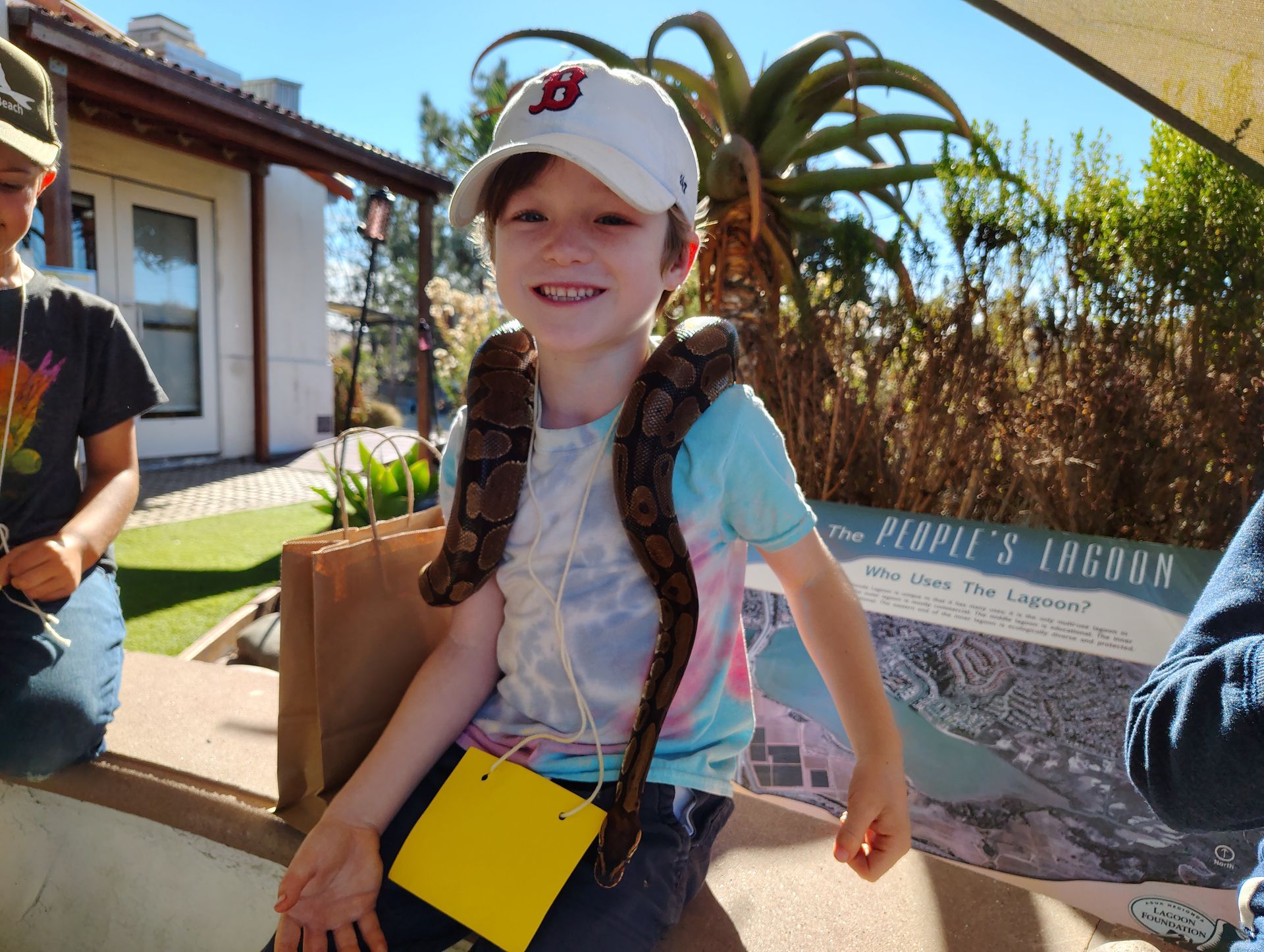 Boy with a snake draped around his neck, smiling. Outdoors, wearing a baseball cap.