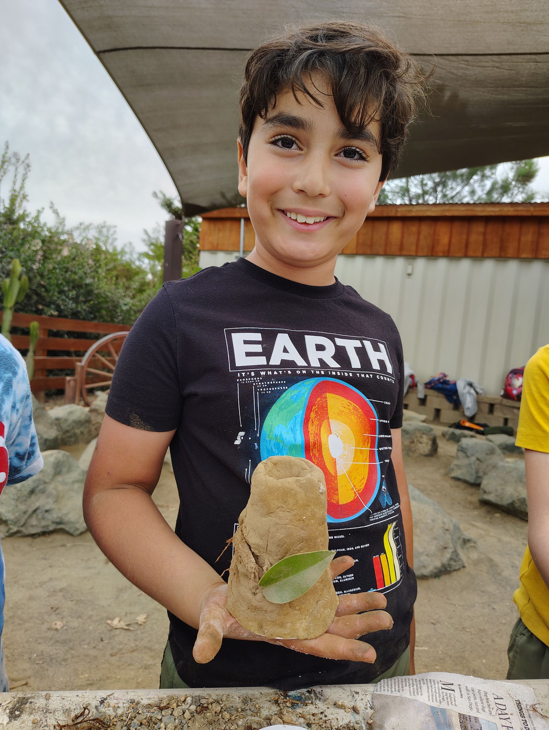 Boy smiling, holding a sand sculpture with a leaf in front of a wood building, wearing an 