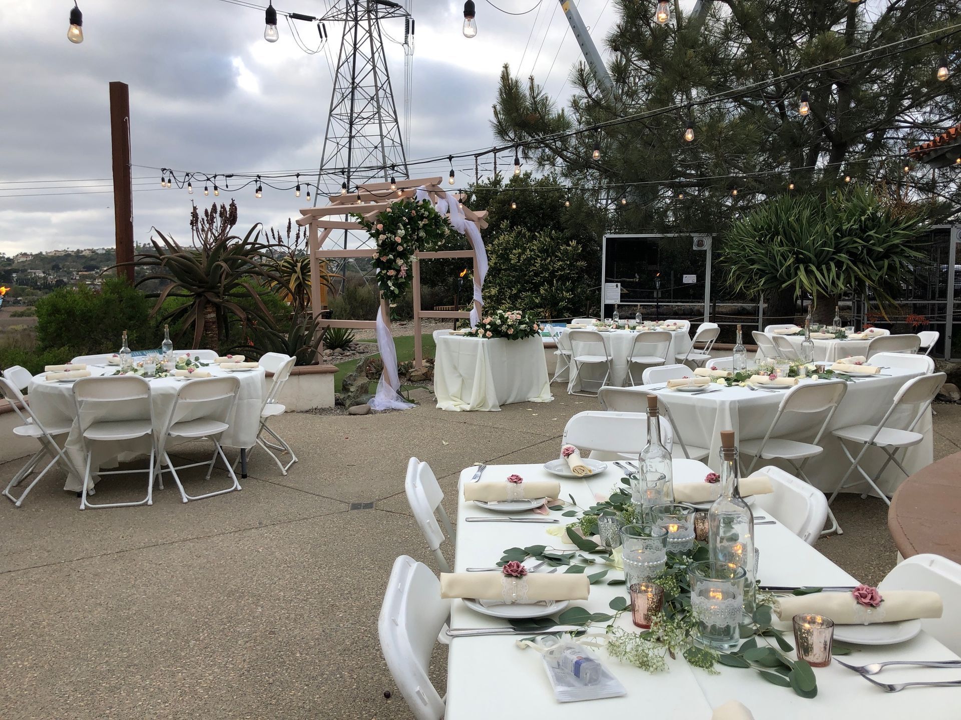 Outdoor event setup with white tables, chairs, and floral centerpieces. String lights and power lines in the background.