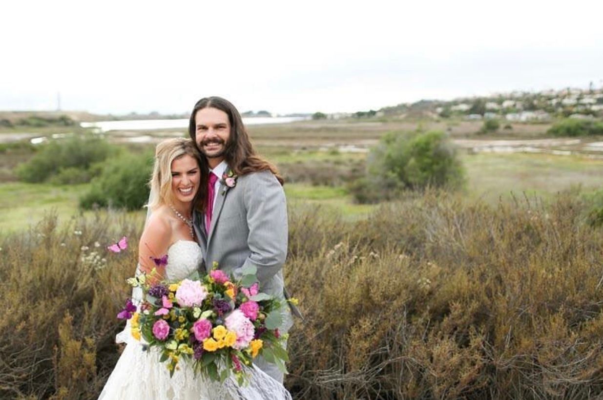 Bride and groom smile, holding bouquet. Cloudy day, grassy field and coastal view in background.
